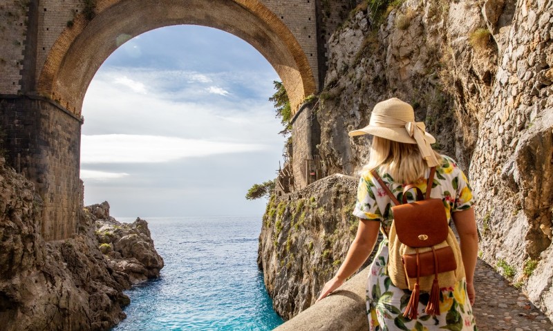 A woman in a floral dress and hat gazes at the serene sea through an ancient stone archway, surrounded by rocky cliffs.