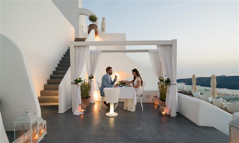 A couple enjoys a romantic dinner on a terrace, surrounded by white curtains and lanterns, with stunning ocean views at sunset.