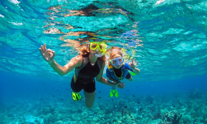 Une femme et un enfant nagent sous l'eau, souriants et saluant de la main, entourés d'une eau bleue éclatante et de récifs coralliens.