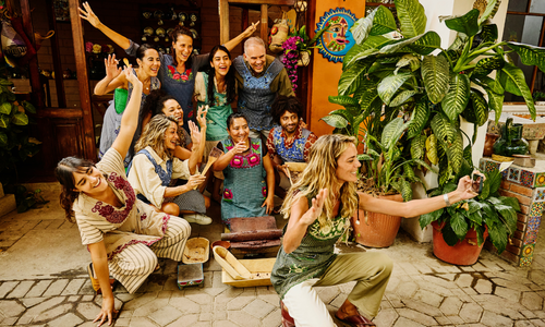 Un groupe de personnes posant et souriant à l'intérieur, dont l'une prend un selfie, dans un décor aux couleurs vives entouré de grandes plantes en pot.