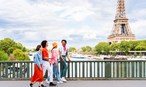 Quatre personnes marchent et discutent sur un pont près de la Tour Eiffel à Paris, avec la Seine et des arbres en arrière-plan.