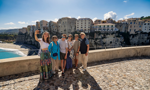 Un groupe de six touristes prenant un selfie par une journée ensoleillée, avec en arrière-plan les falaises du littoral et des bâtiments historiques.
