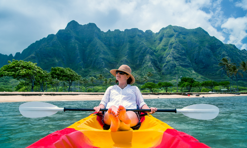 Une femme se détend dans un kayak sur une eau calme, avec en arrière-plan des montagnes verdoyantes et un ciel bleu.