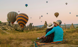 Couple sitting on a colorful rug in a field, watching multiple hot air balloons float over rocky terrain at sunset.